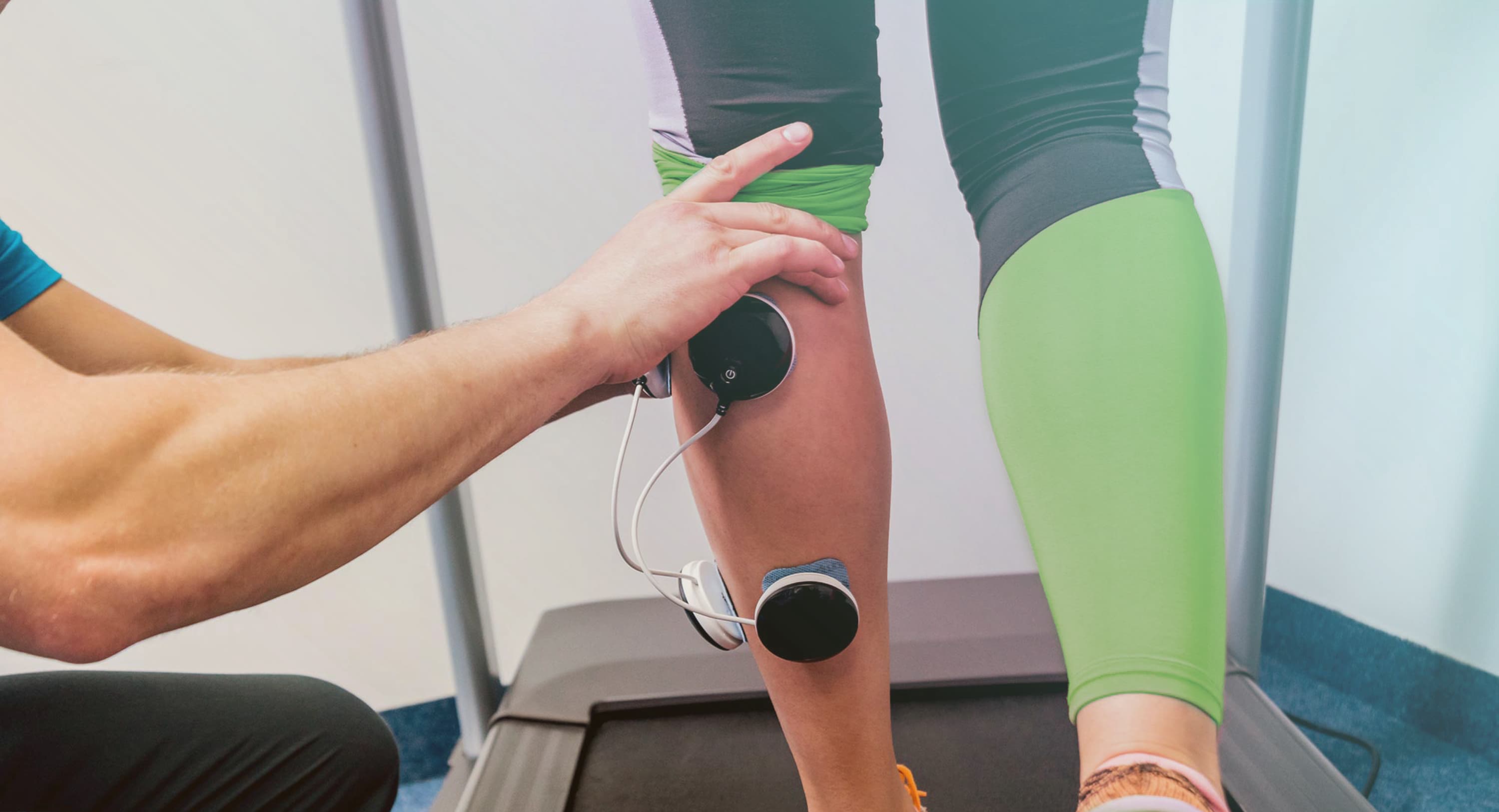 Patiënt op een loopband, fysiotherapeut plakt elektroden op kuiten van patiënt.