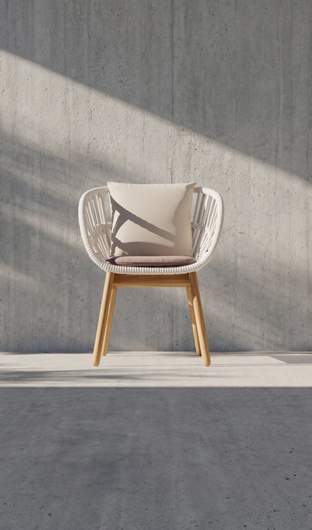 A modern chair with a wooden frame and white woven seat, topped with a light-colored cushion, sits against a gray concrete wall with sunlight casting diagonal shadows across the scene.