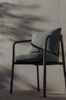 A modern chair with a green cushion and pillow sits against a concrete wall, with shadows of leaves and branches cast by sunlight across the wall and chair.