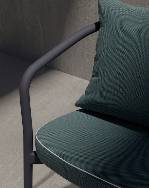 A close-up of a modern dark green cushioned chair with a sleek black metal armrest, positioned against a gray concrete wall in soft natural light.