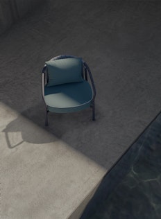 A single modern blue chair with a matching cushion sits on a concrete floor near the edge of a pool. Strong sunlight casts a shadow of the chair and creates contrasting light and dark areas on the surface.