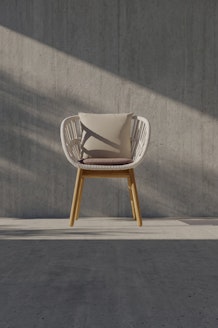A modern chair with a wooden frame and white woven seat, topped with a light-colored cushion, sits against a gray concrete wall with sunlight casting diagonal shadows across the scene.