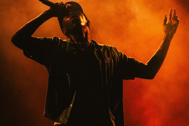 Tyler, The Creator sings into microphone arms raised in front of an amber haze.