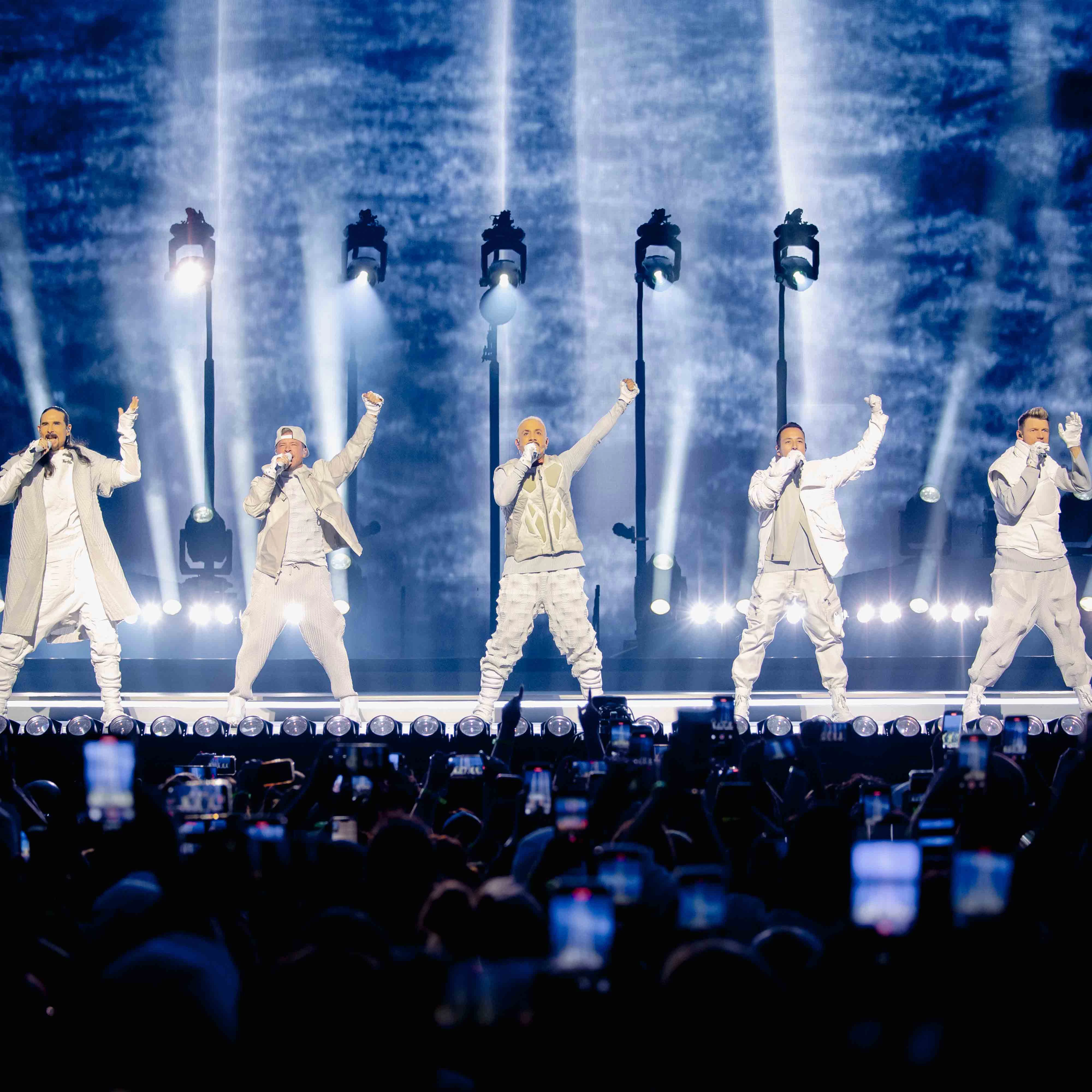 Backstreet Boys on Sphere stage with arms raised under blue and white lighting, audience phones visible in foreground.