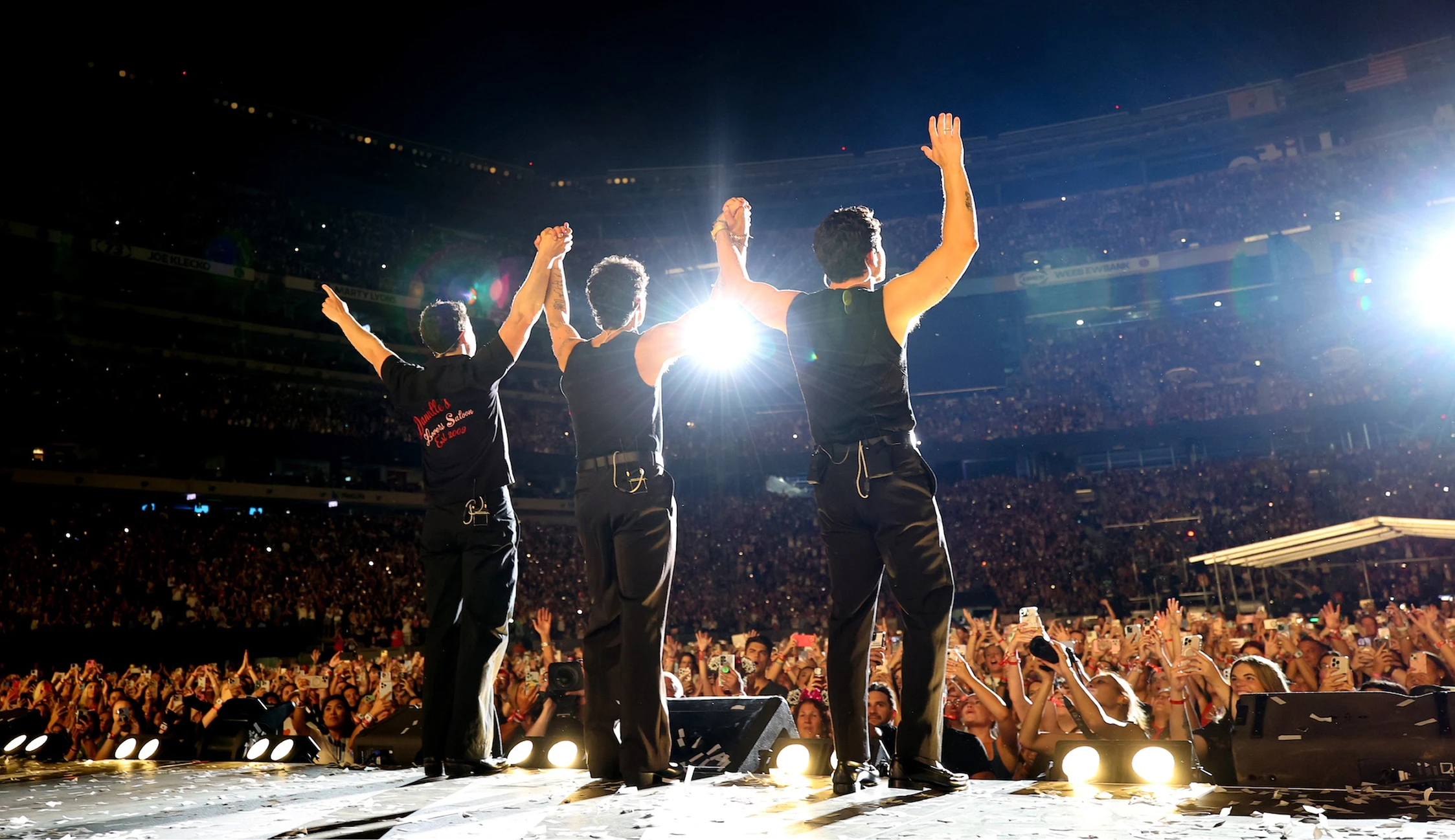 Jonas Brothers stand on stage holding hands arms up in the air facing the crowd