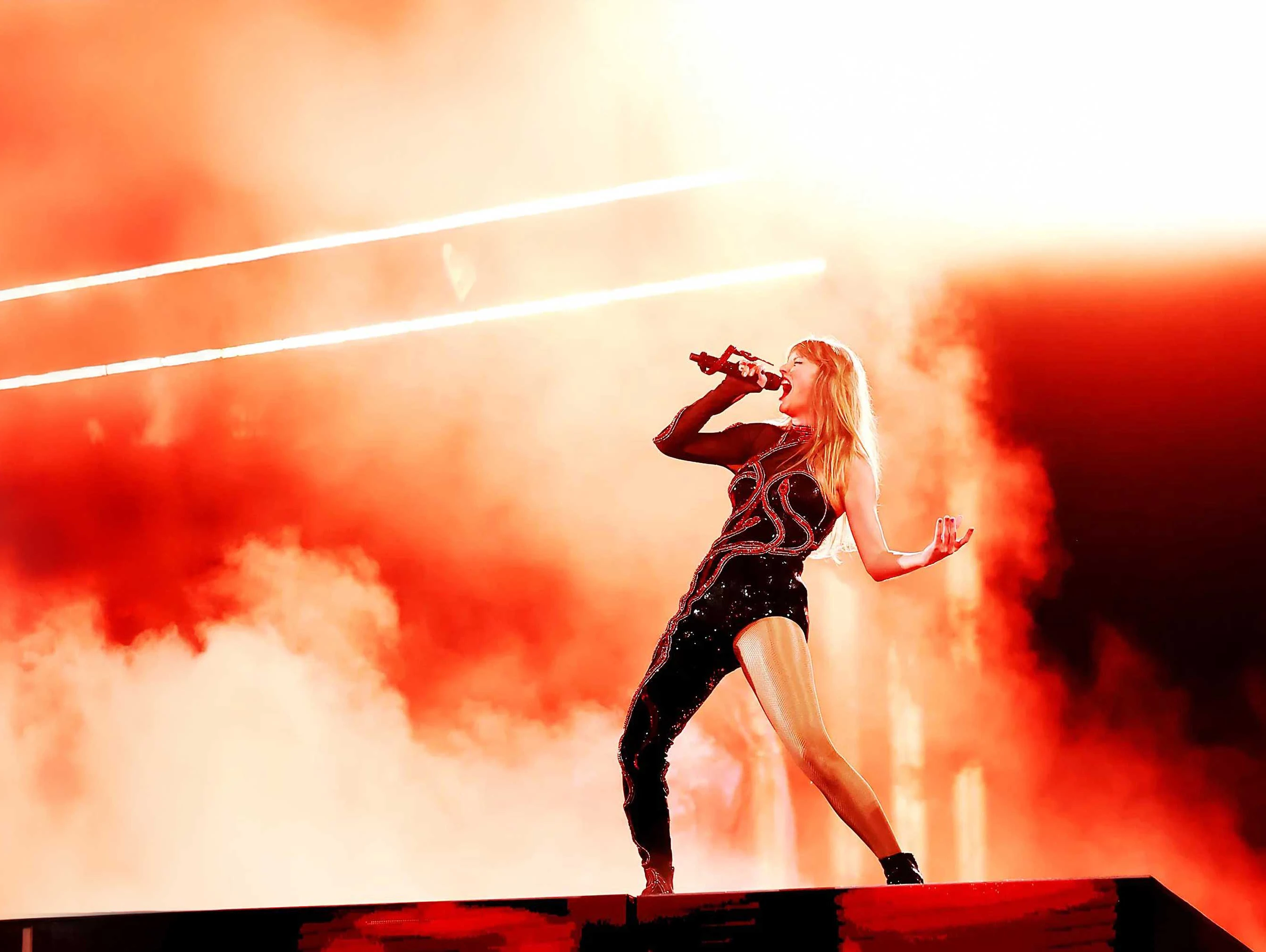 Taylor Swift - Eras Tour - Silent House Taylor Swift sings into microphone in a strong pose staged with fog and amber light
