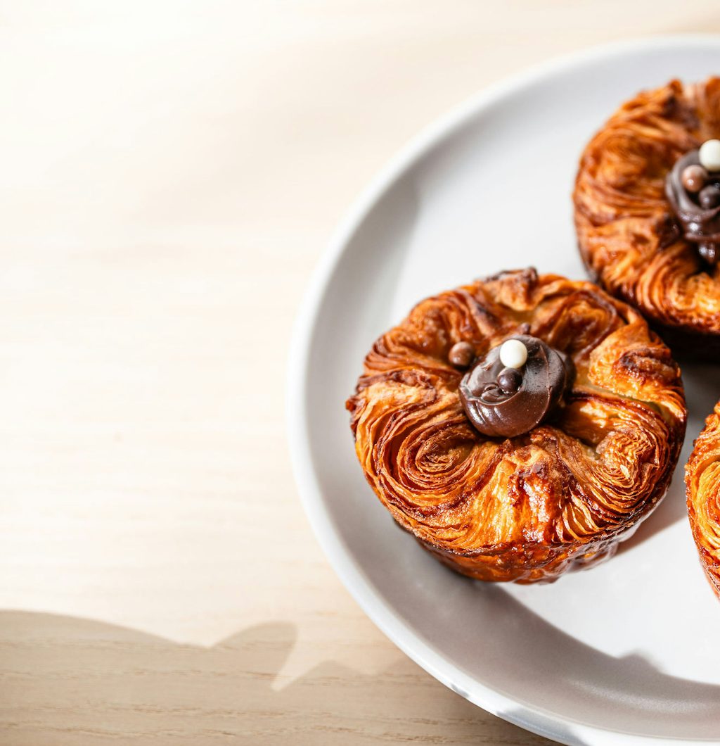 Three round, golden-brown laminated pastries arranged on a white plate, each topped in the center with chocolate and small decorative chocolate pieces.