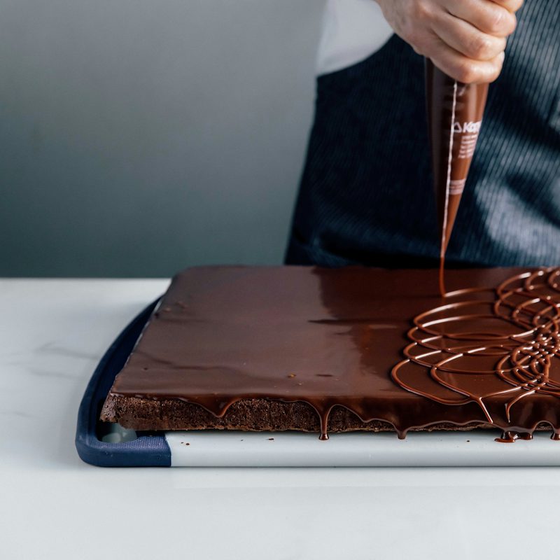 Close-up of a chocolate-covered rectangular dessert being decorated with an intricate floral pattern of piped chocolate on top.