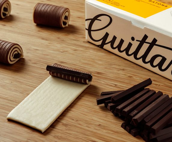 Guittard Chocolate Batons packaging displayed on a wooden surface alongside dark chocolate batons, rolled chocolate pastry decorations, and a sheet of pastry dough partially wrapped around a chocolate baton, illustrating baking and pastry applications.