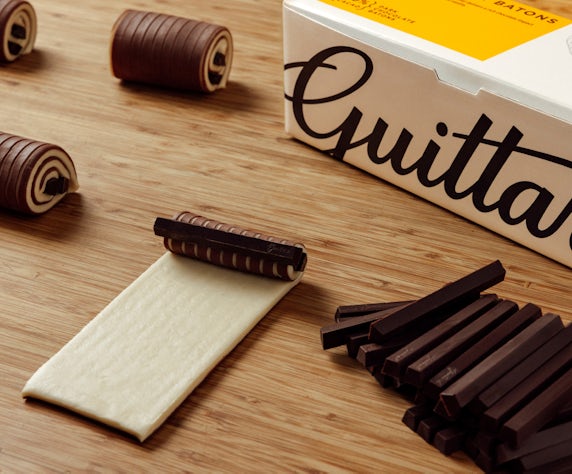 Guittard Chocolate Batons packaging displayed on a wooden surface alongside dark chocolate batons, rolled chocolate pastry decorations, and a sheet of pastry dough partially wrapped around a chocolate baton, illustrating baking and pastry applications.
