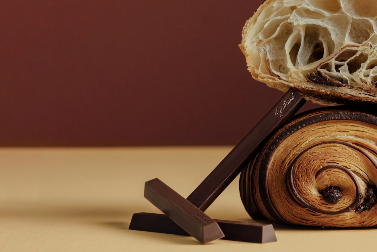 Close-up of a chocolate pastry cut in half, showing an airy, layered interior with large open pockets. The cut pastry rests on top of a rolled chocolate pastry, with three dark chocolate batons arranged in front on a tan surface against a brown background.