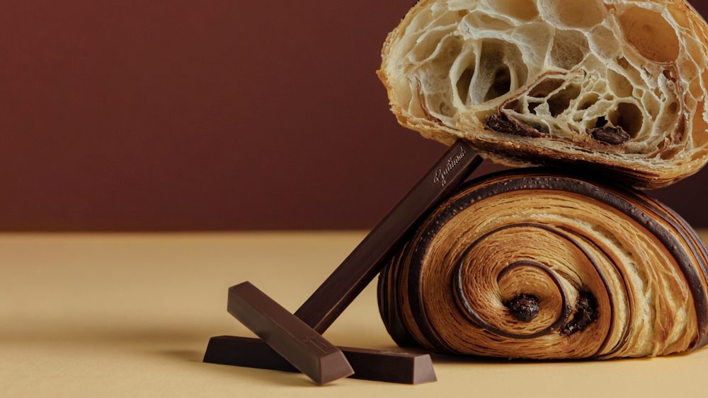Close-up of a chocolate pastry cut in half, showing an airy, layered interior with large open pockets. The cut pastry rests on top of a rolled chocolate pastry, with three dark chocolate batons arranged in front on a tan surface against a brown background.