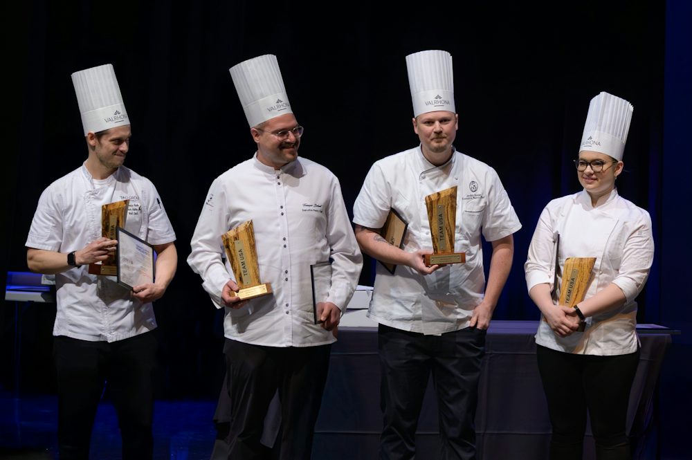 Four pastry chefs wearing white chef jackets and tall chef hats stand on a stage holding awards and certificates during a culinary competition or recognition ceremony.