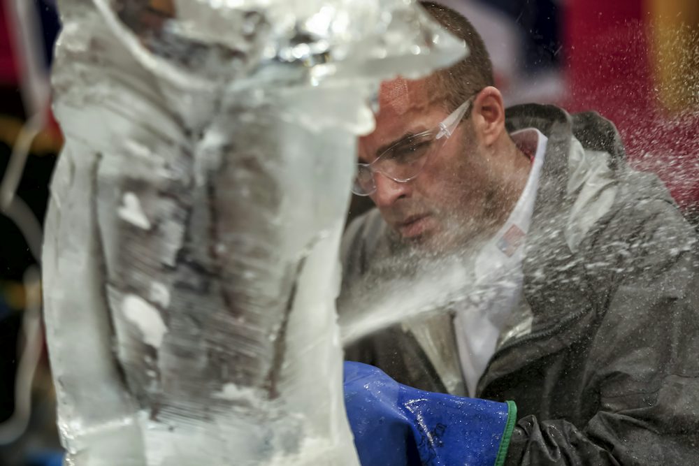 Person wearing safety glasses and protective gloves carving an ice sculpture, with ice chips and water spray visible around the work area during a live ice sculpting demonstration.