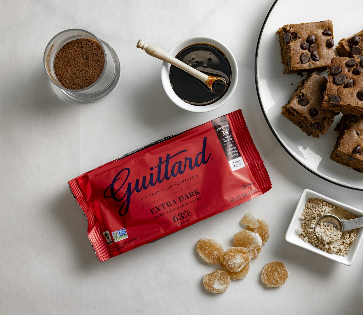 Overhead view of chocolate chip cookie bars beside a red Guittard Extra Dark chocolate bar, cocoa powder, ginger pieces, and baking ingredients.