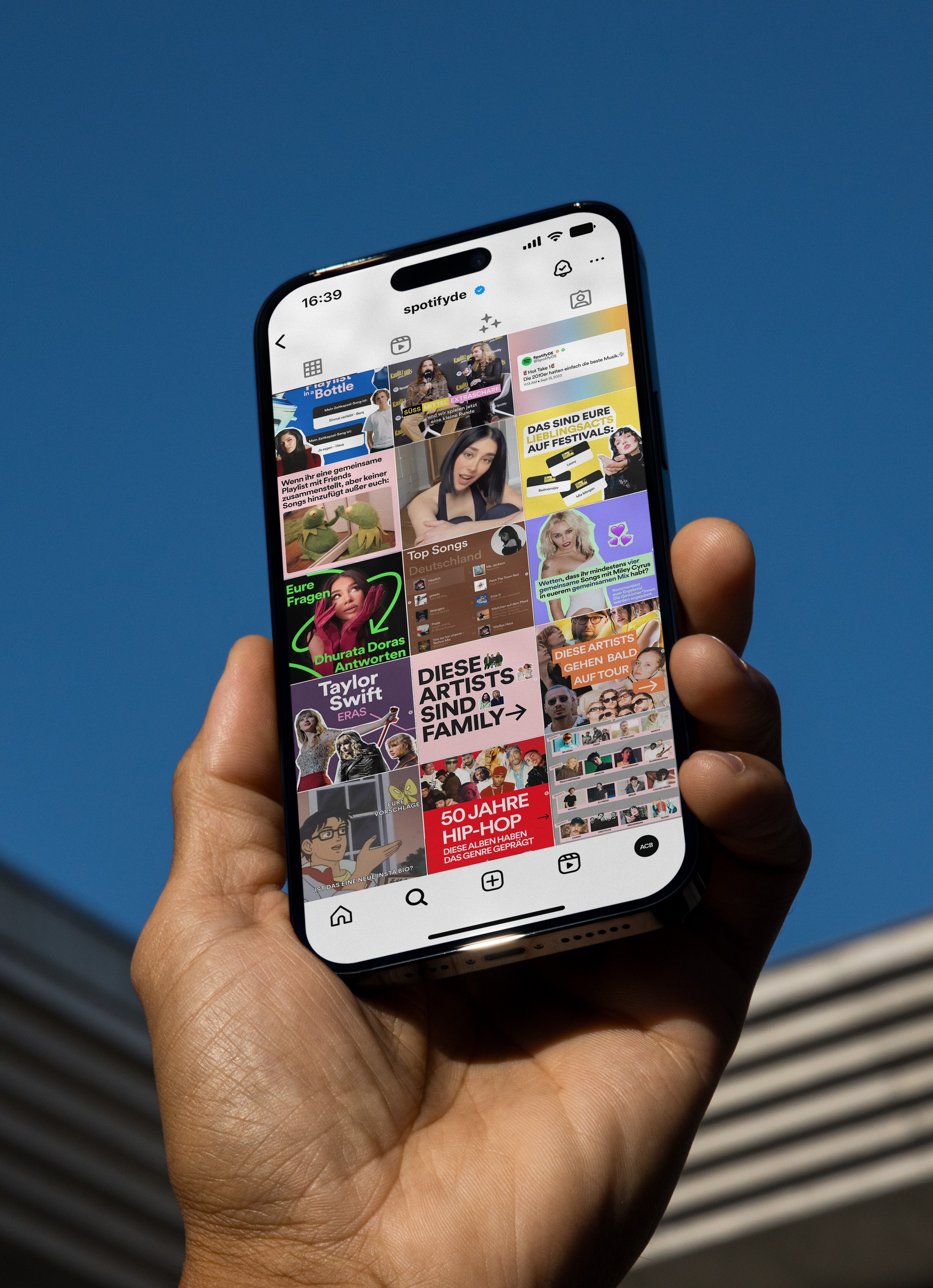 A hand holding a smartphone displaying an Instagram feed from the user ‘spotifyde’. The feed features various colorful posts, including images and text related to music and artists, such as Taylor Swift, Dhurata Dora, and mentions of hip-hop and festival activities. The background is a clear blue sky.