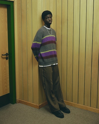 A young man wearing a full carhartt outfit standing against a wooden paneled wall. He is wearing a gray sweater with horizontal purple and green stripes, paired with brown trousers and black shoes. He has a short afro hairstyle and a thin silver chain around his neck.
