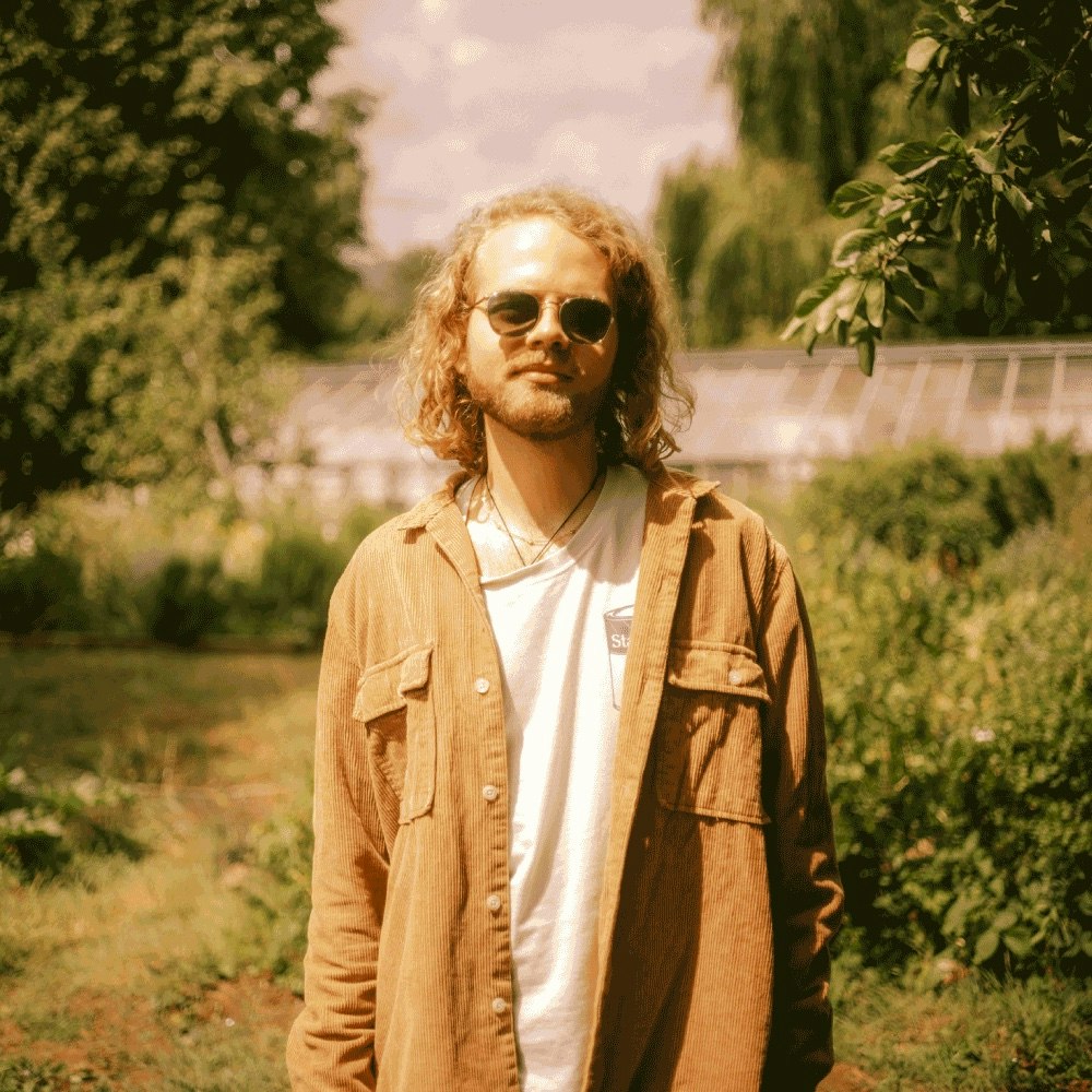 This image may contain: coat, jacket, person, photography, portrait, sunglasses, tree, grass, vegetation, smile