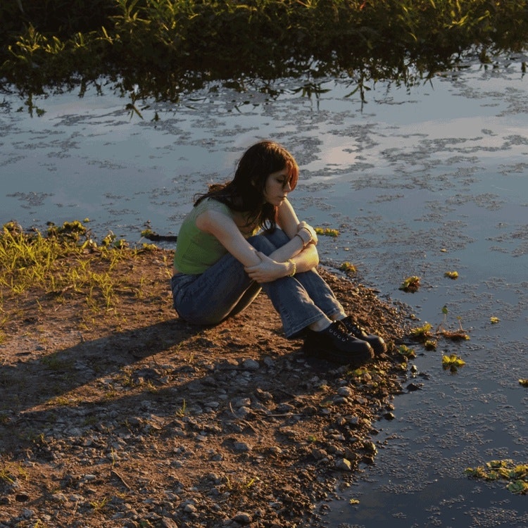 This image may contain: person, sitting, pants, land, photography, female, girl, teen, portrait, gravel