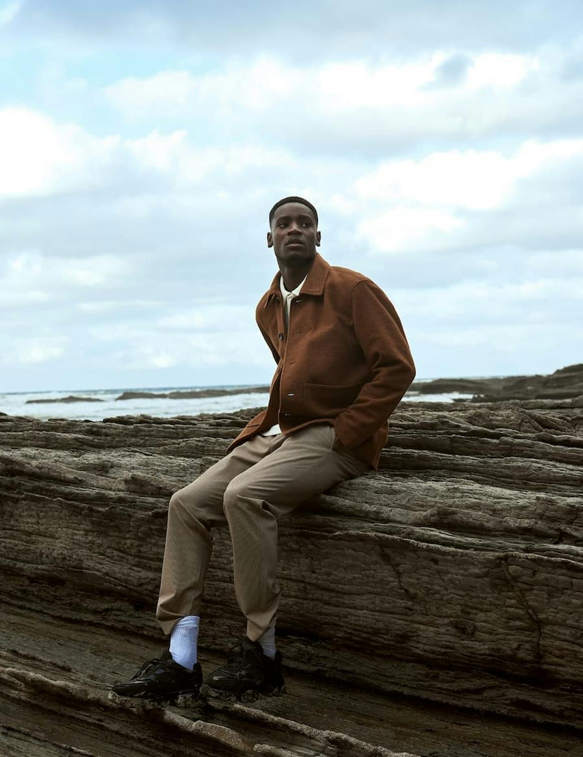 Well dressed man sitting on a rocky shore, looking inland, hands in pockets, slightly hunched forward