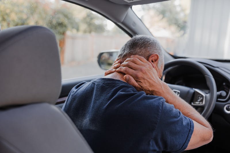 man behind the wheel of a car holding the back of his neck with both hands