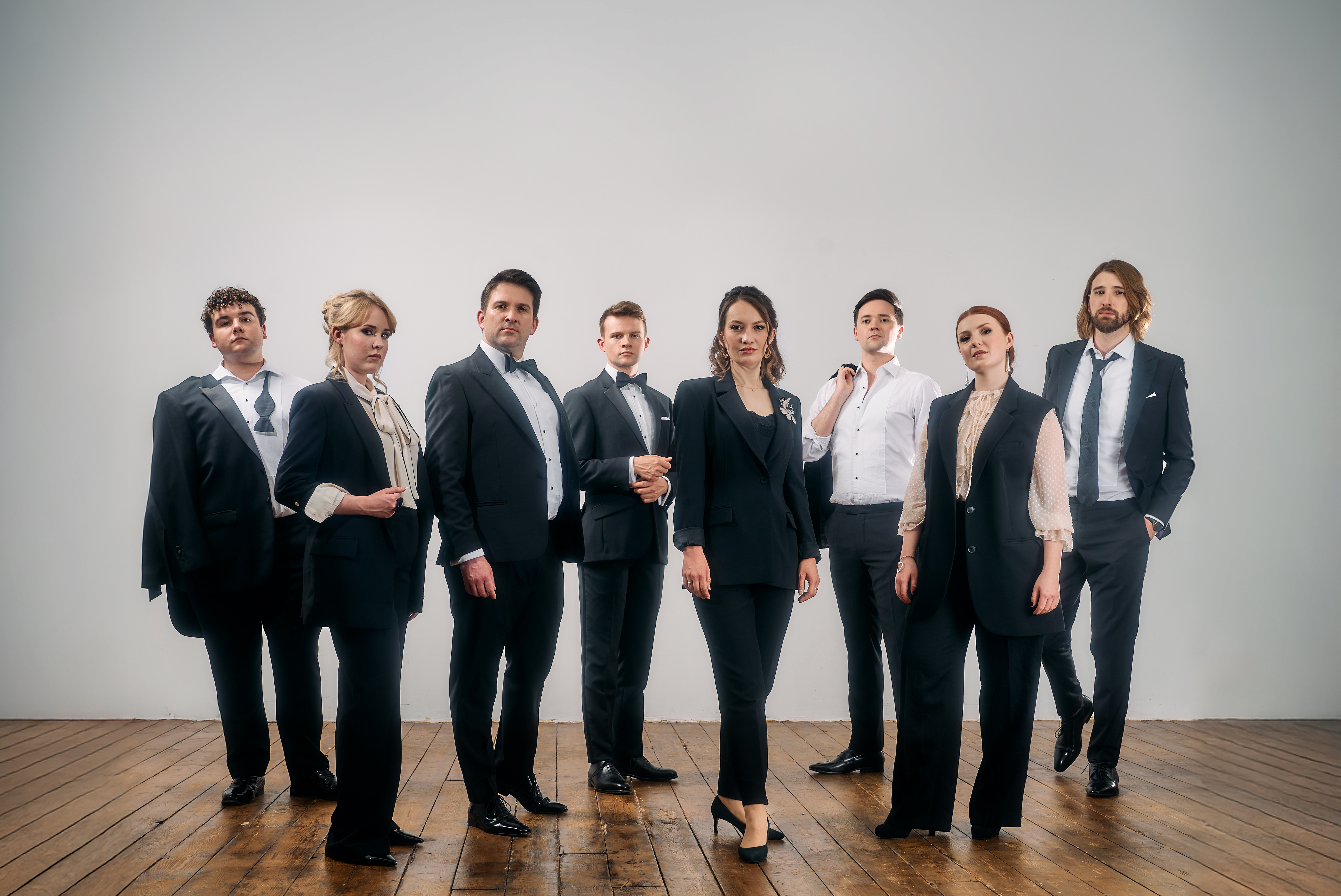 A vocal ensemble of 8 people stand together posing on a dark hardwood floor