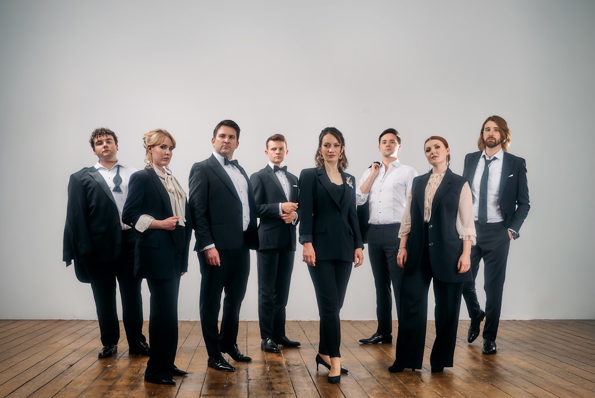 A vocal ensemble of 8 people stand together posing on a dark hardwood floor