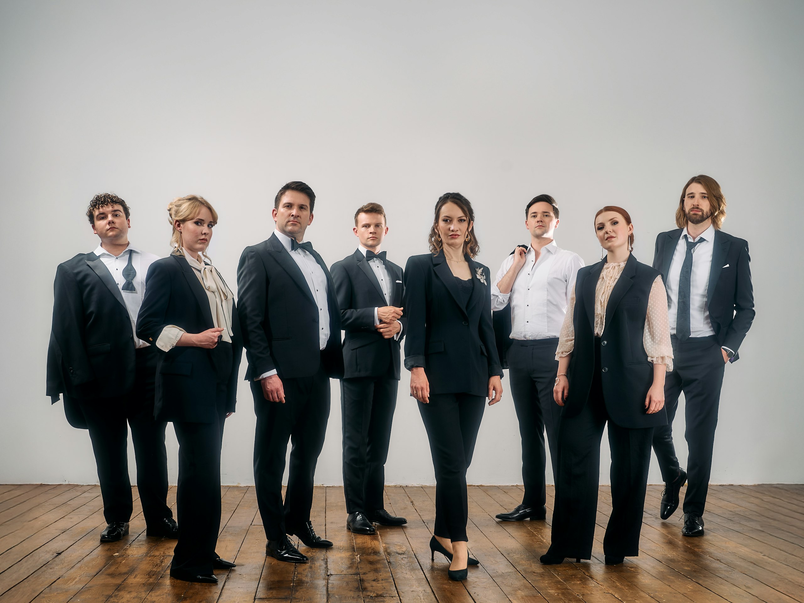 A vocal ensemble of 8 people stand together posing on a dark hardwood floor