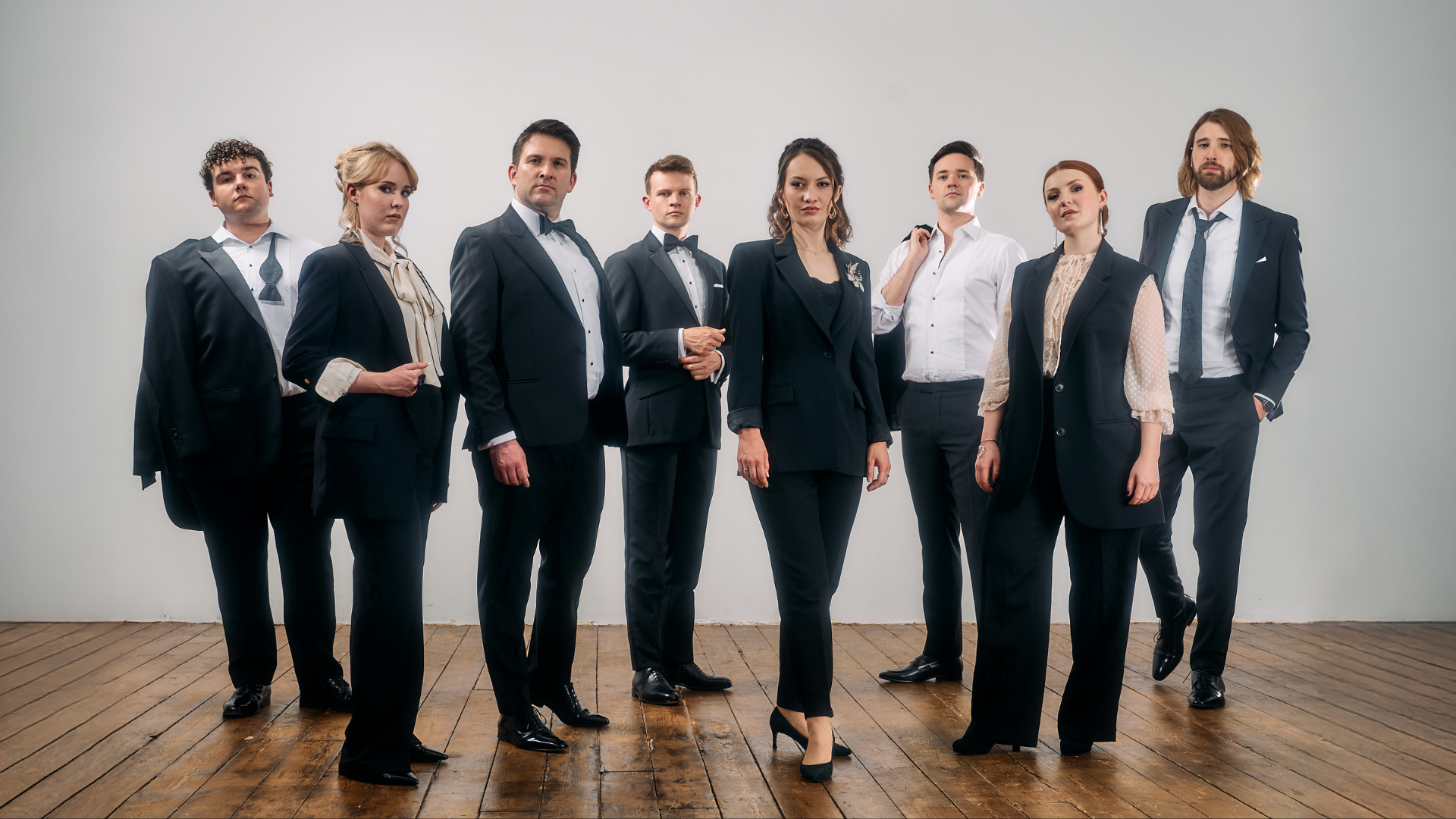 A group of 8 members of a vocal ensemble stand together in vests and suits.