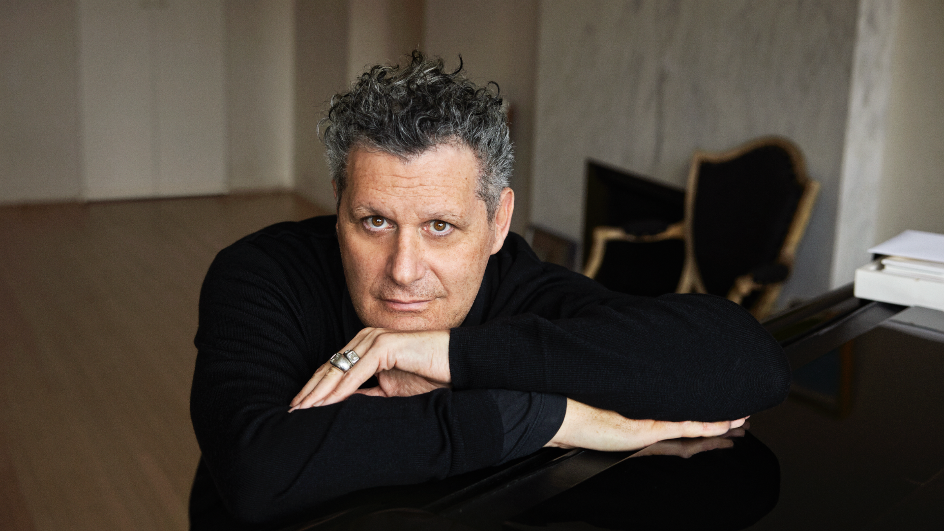 Isaac Mizrahi leans forward on his piano, looking into the camera.