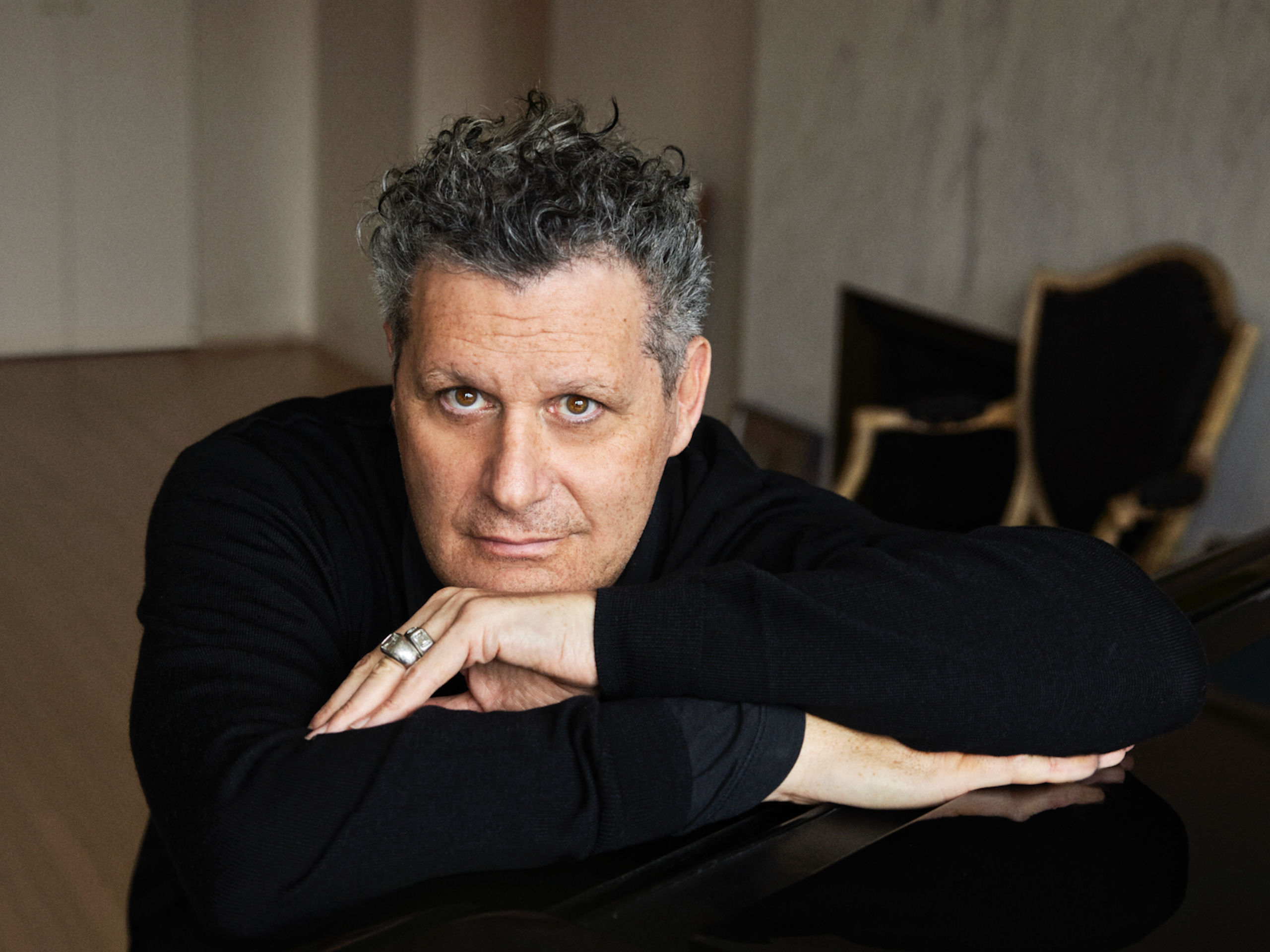 Isaac Mizrahi leans forward on his piano, looking into the camera.