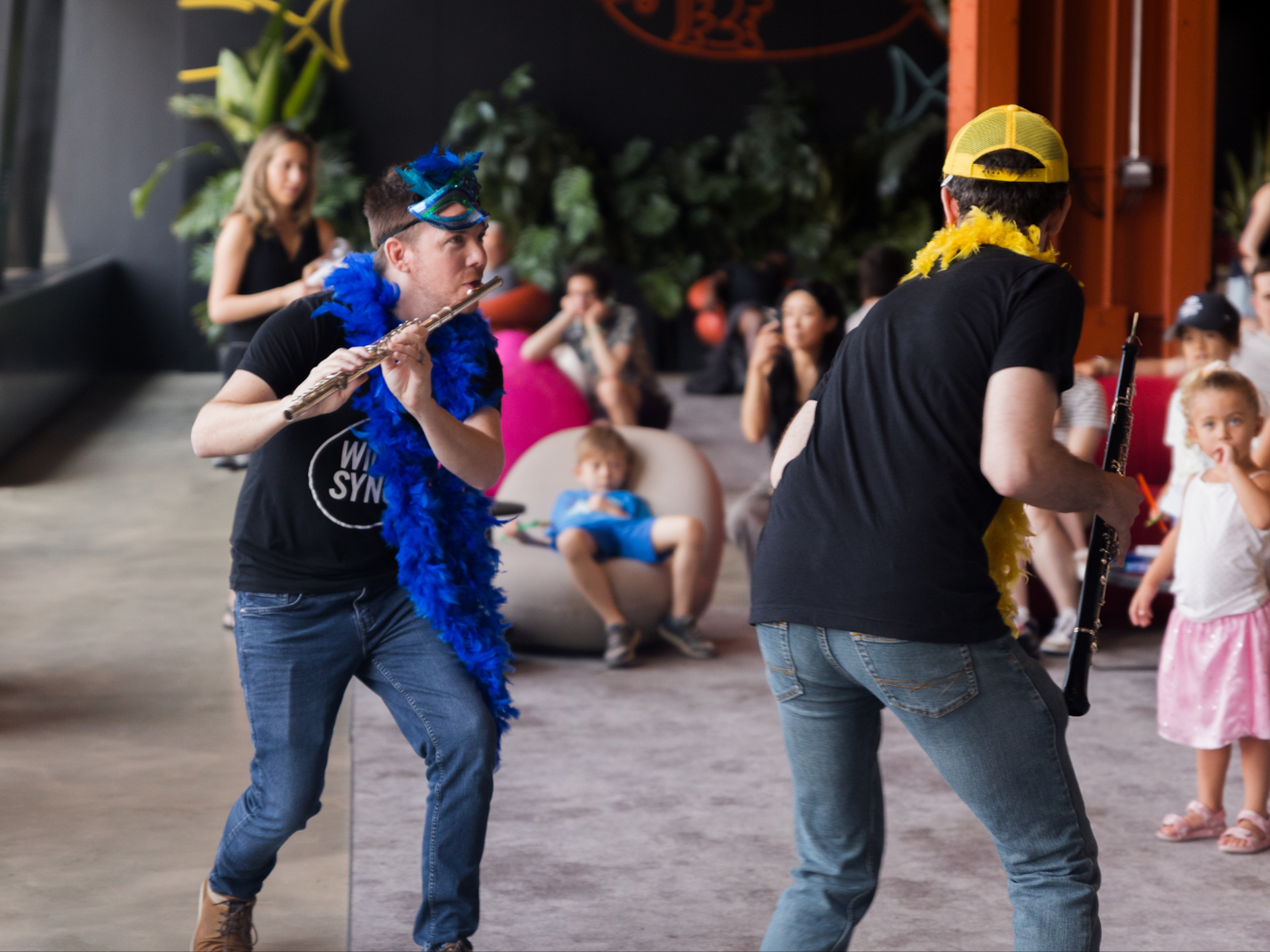 A flutist dressed with a boa and a mask performs in front of onlooking children.