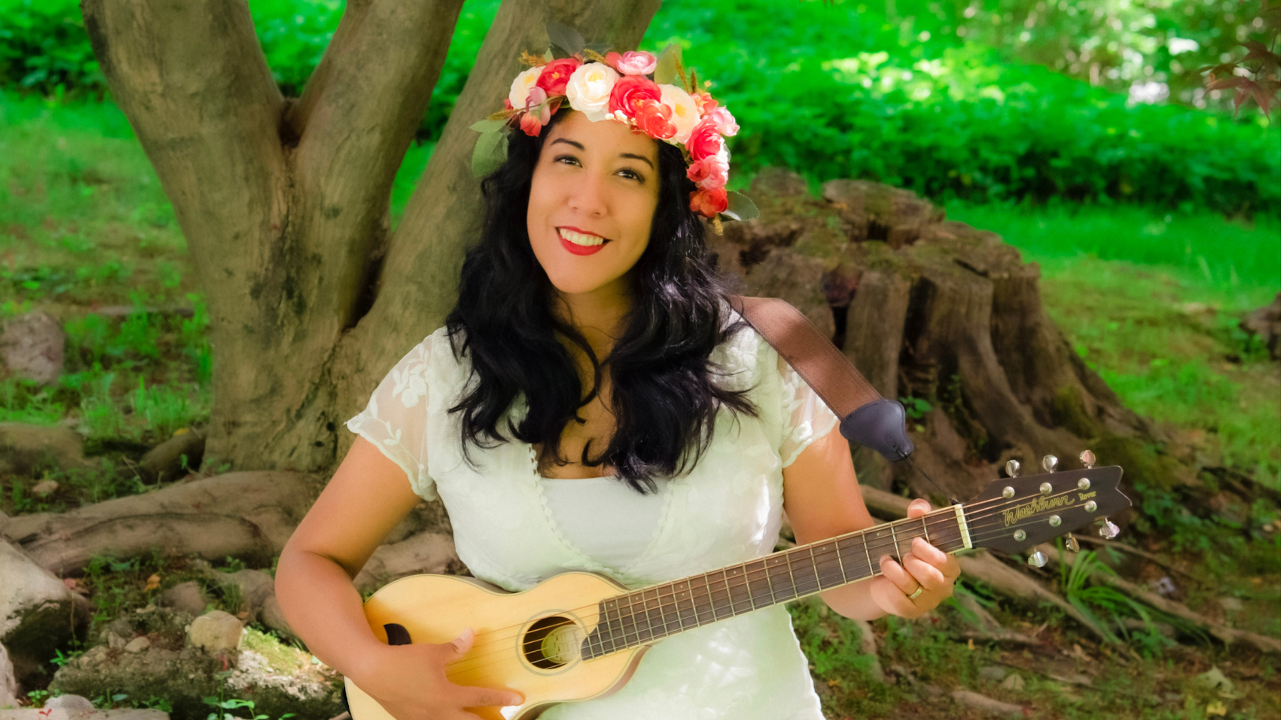 A woman in a flower crown holds a little guitar in a wooded setting.