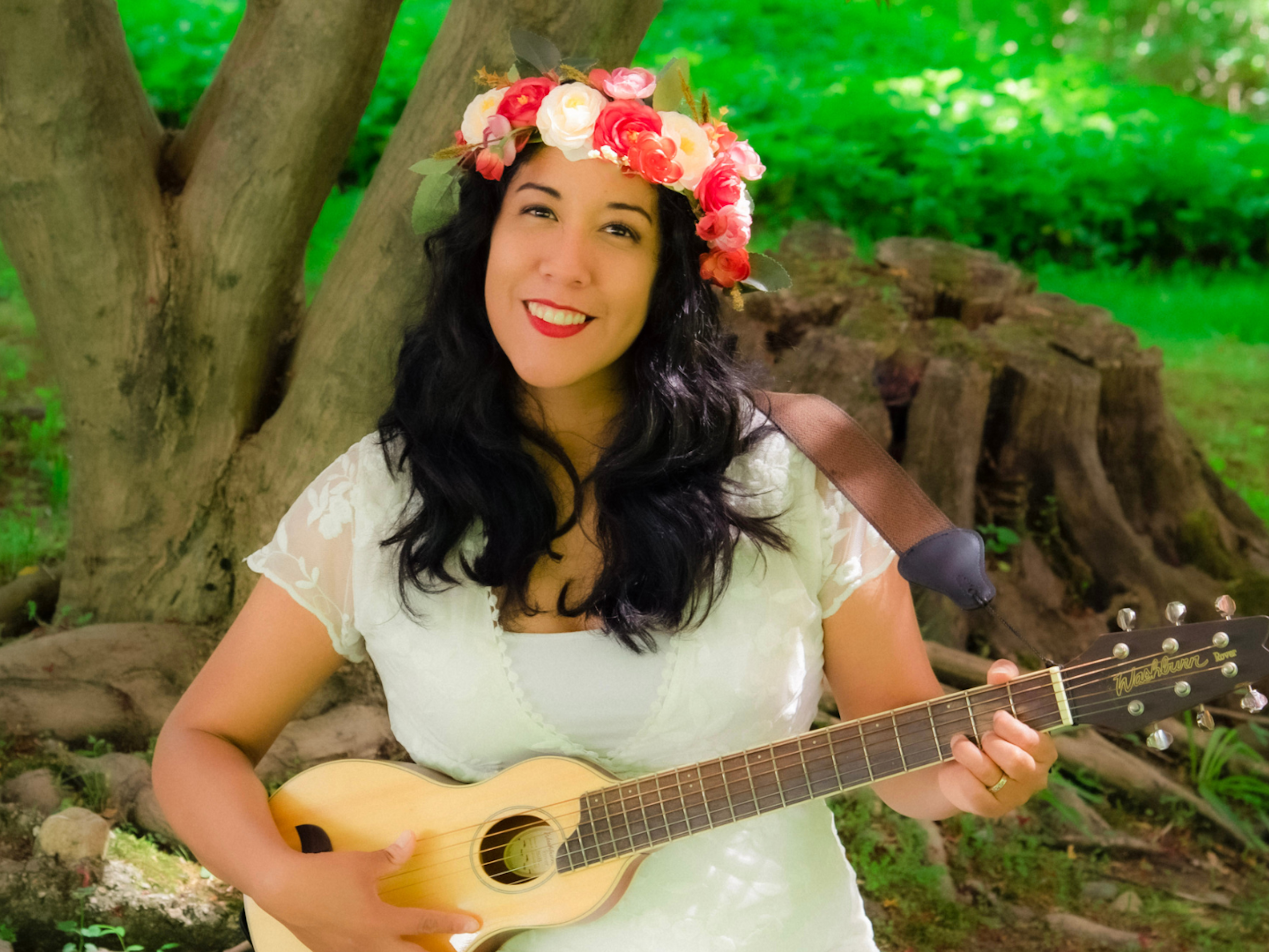 A woman in a flower crown holds a little guitar in a wooded setting.