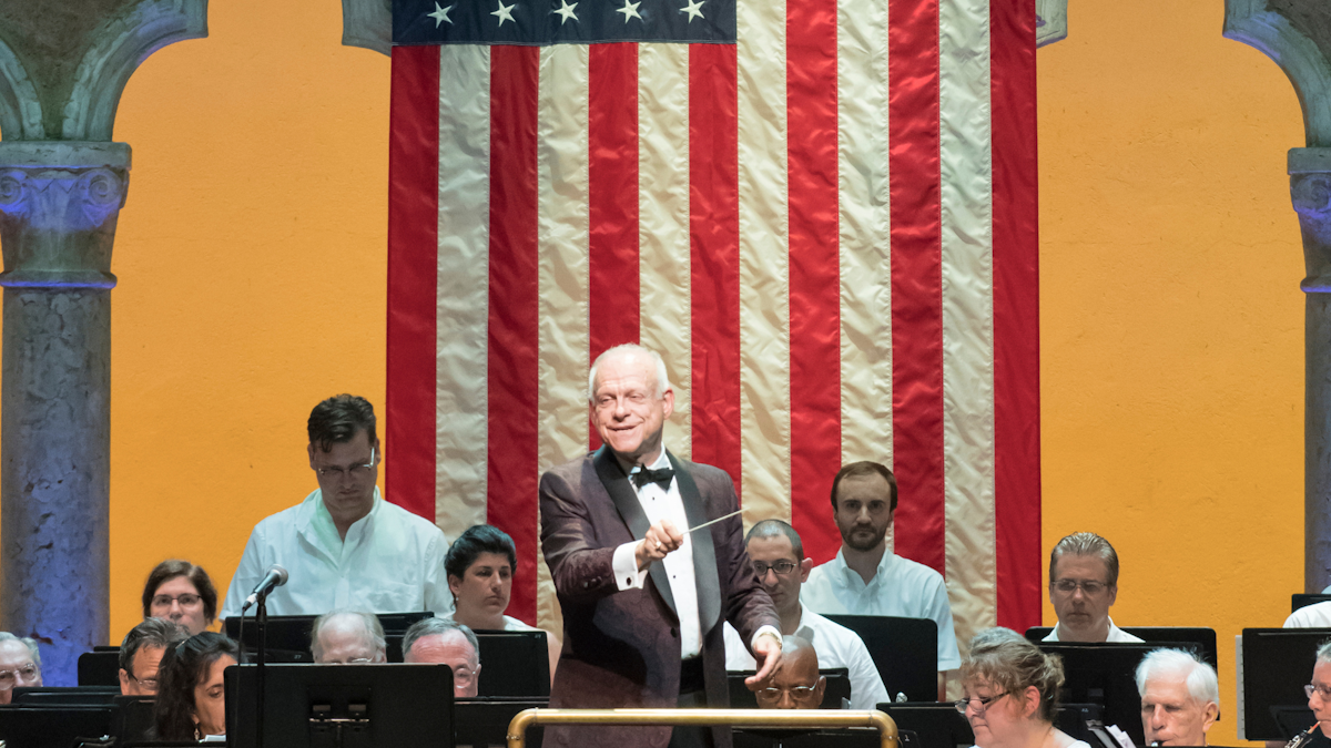 A conductor in a tuxedo waves his baton in the Venetian Theater. There is an orchestra playing behind him and an American Flag mounted on the back of the stage.