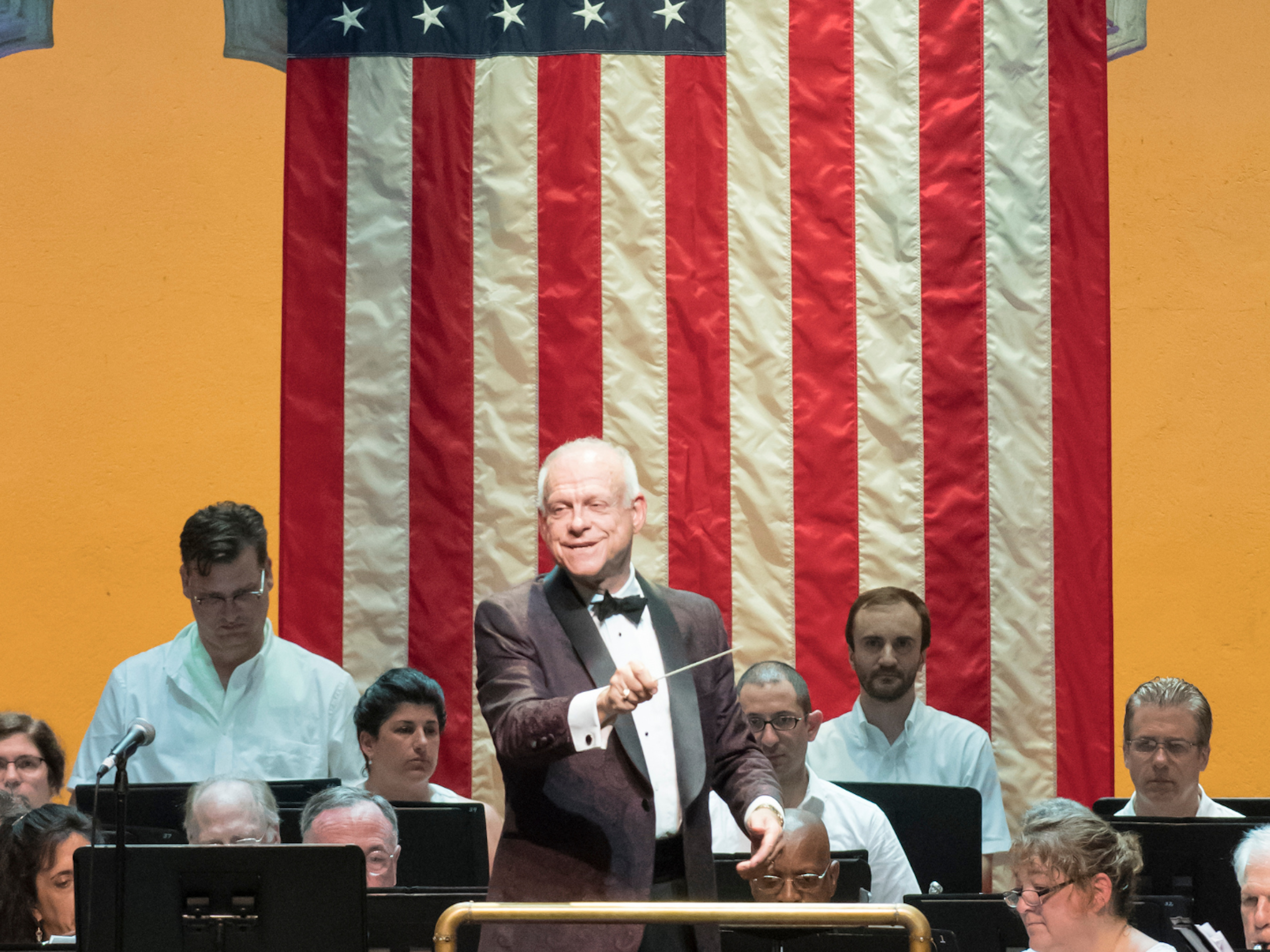 A conductor in a tuxedo waves his baton in the Venetian Theater. There is an orchestra playing behind him and an American Flag mounted on the back of the stage.