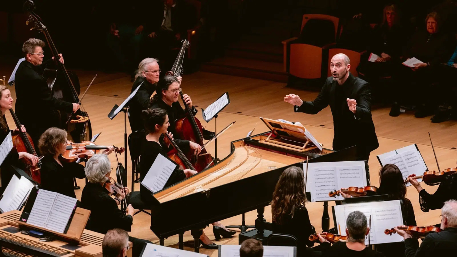 A man conducts a baroque orchestra. Orchestra members on stage play string instruments and the harpsichord.