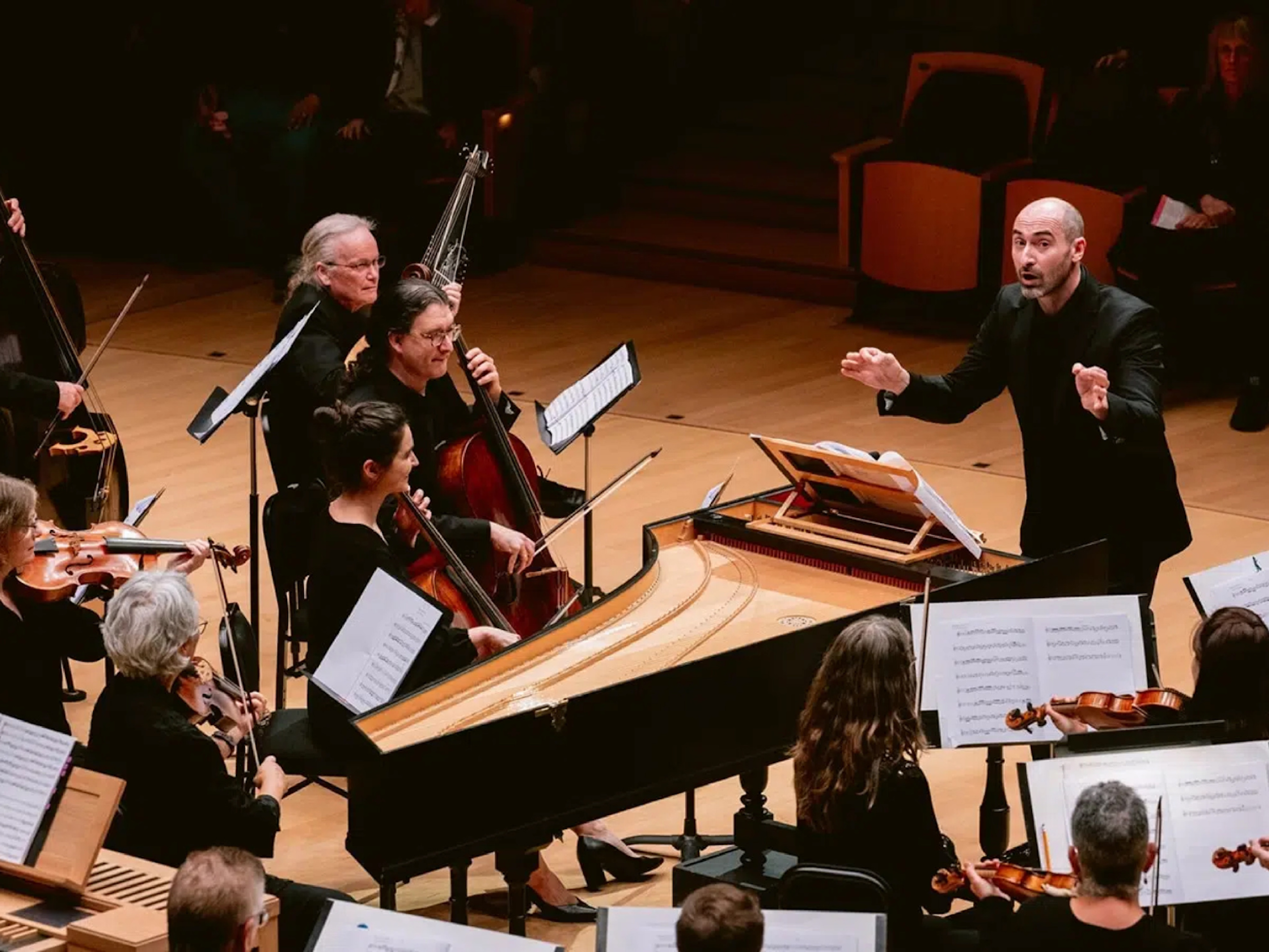 A man conducts a baroque orchestra. Orchestra members on stage play string instruments and the harpsichord.