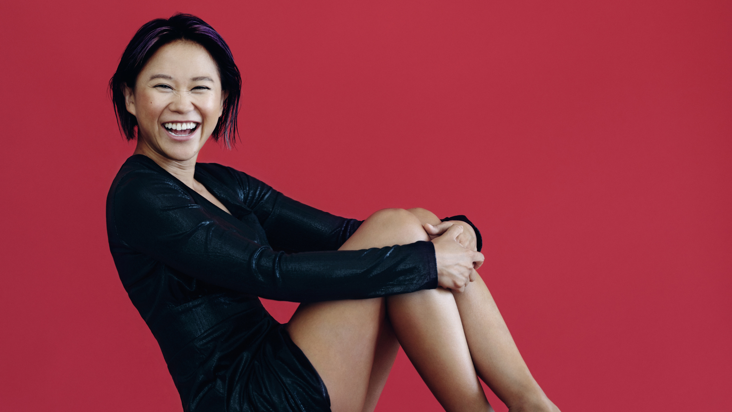 A woman smiles widely while sitting on a chair and hugging her knees.