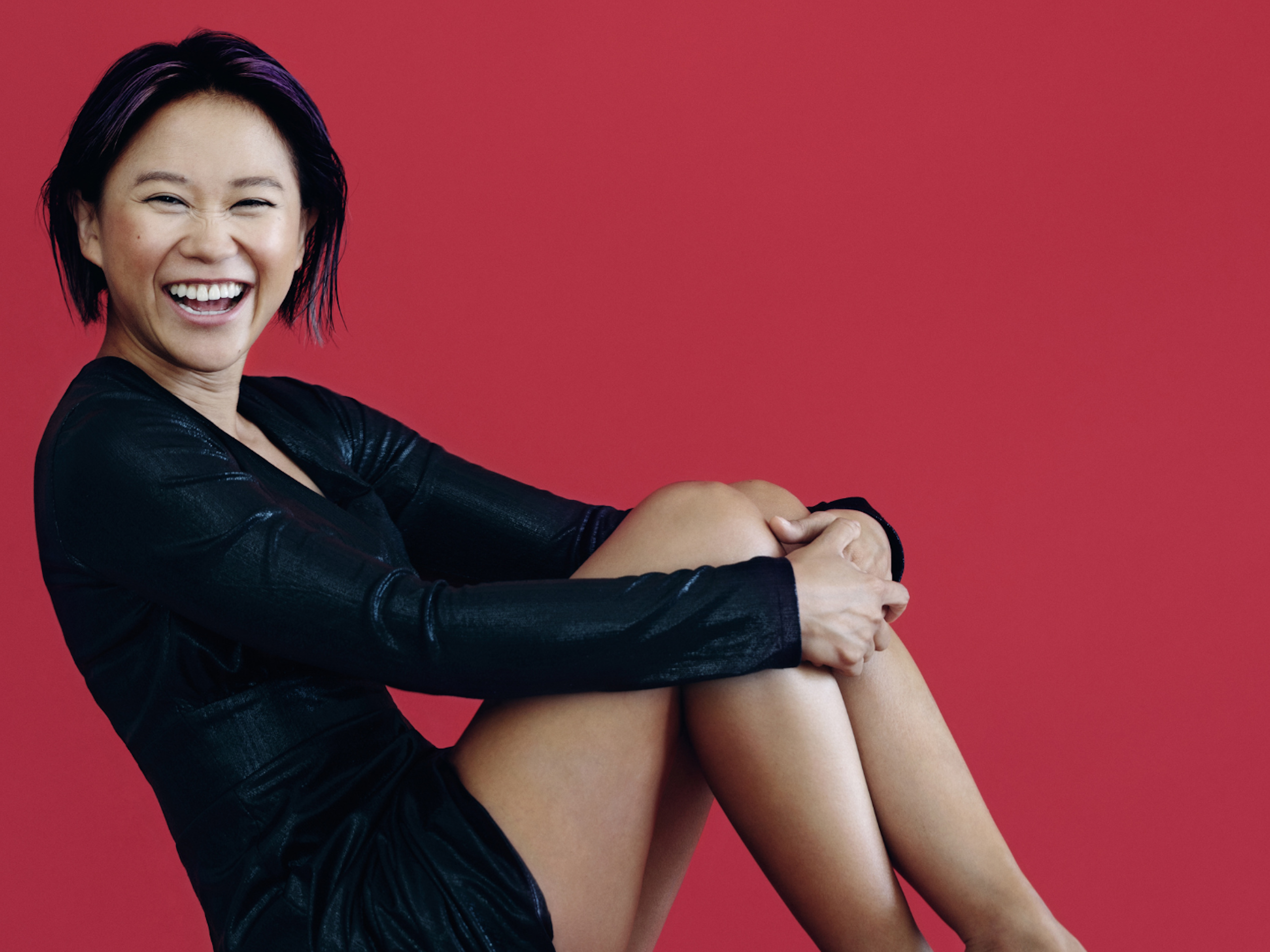A woman smiles widely while sitting on a chair and hugging her knees.