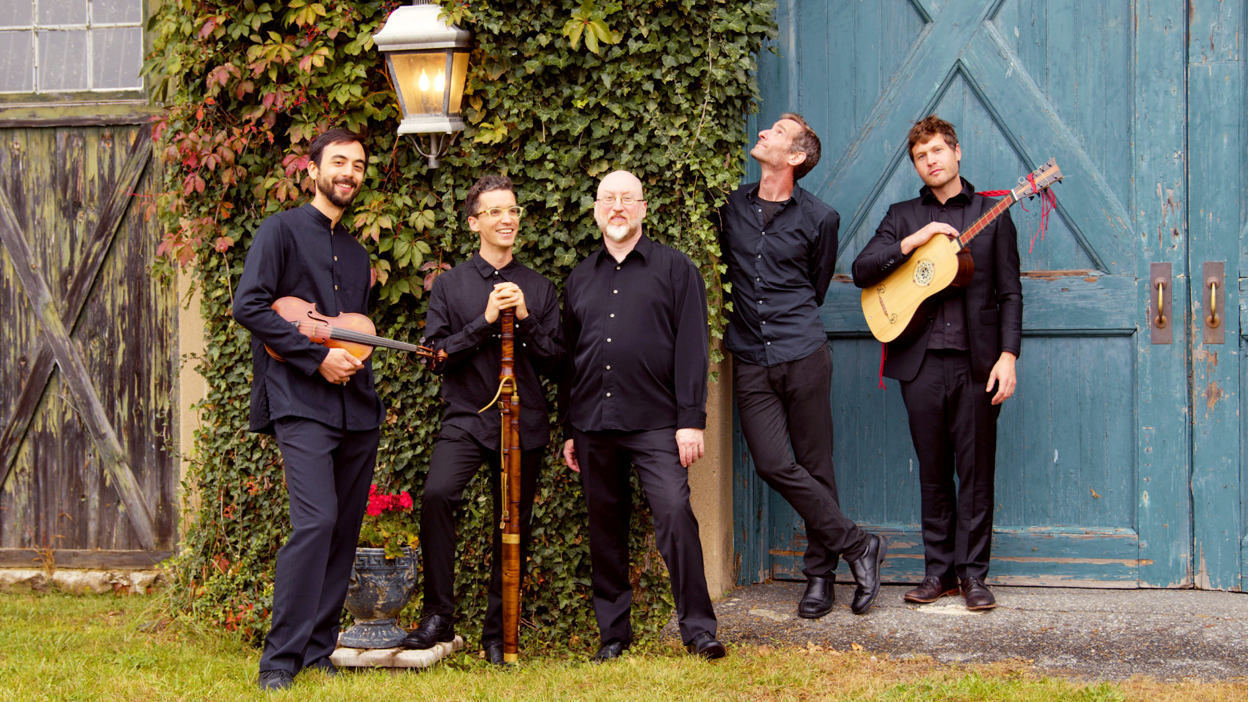 A group of musicians stand together holding their instruments in front of an old barn covered in vines.