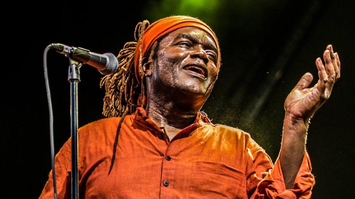 A close up shot of a man at a microphone. There is a green spotlight illuminating him on a dark stage.