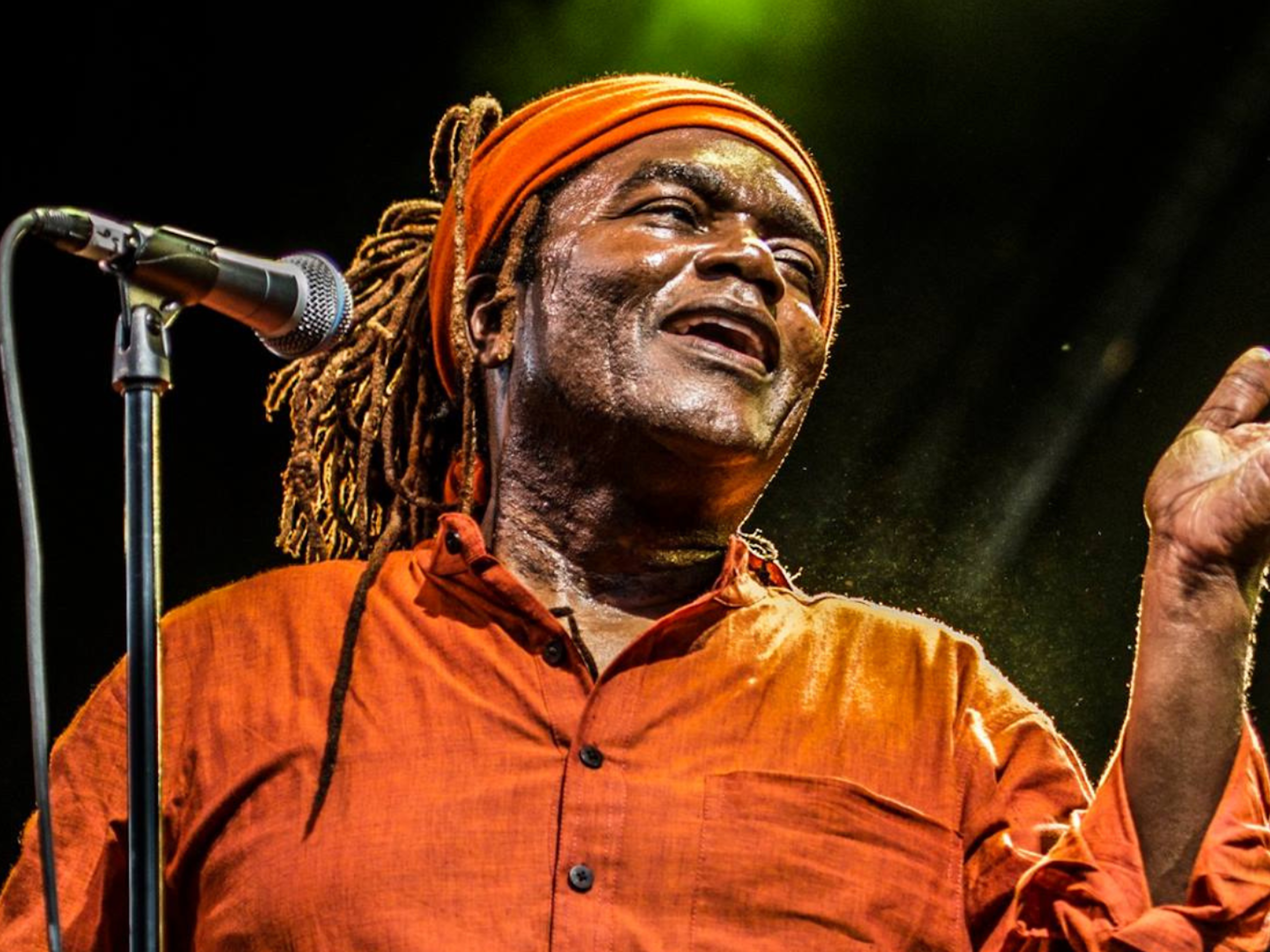 A close up shot of a man at a microphone. There is a green spotlight illuminating him on a dark stage.