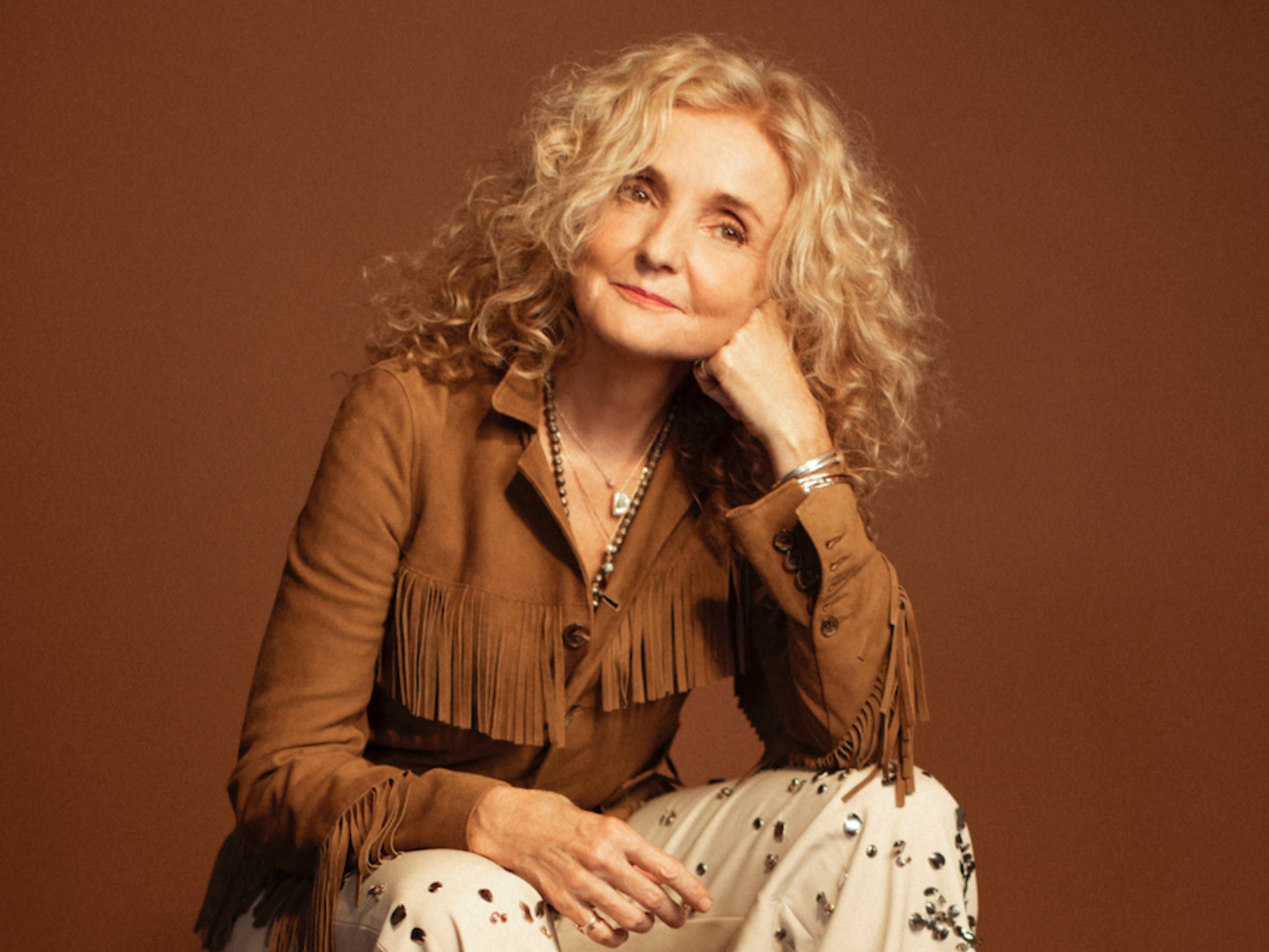 A woman in a fringe blouse sits forward on a box.