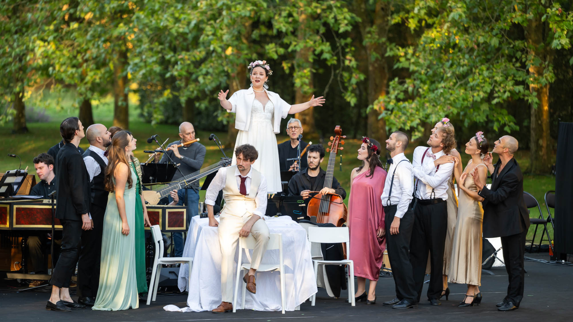 A baroque opera group performs an opera in a lush outdoor setting. A woman stands on a table singing while the opera group, in costume, look up at her.