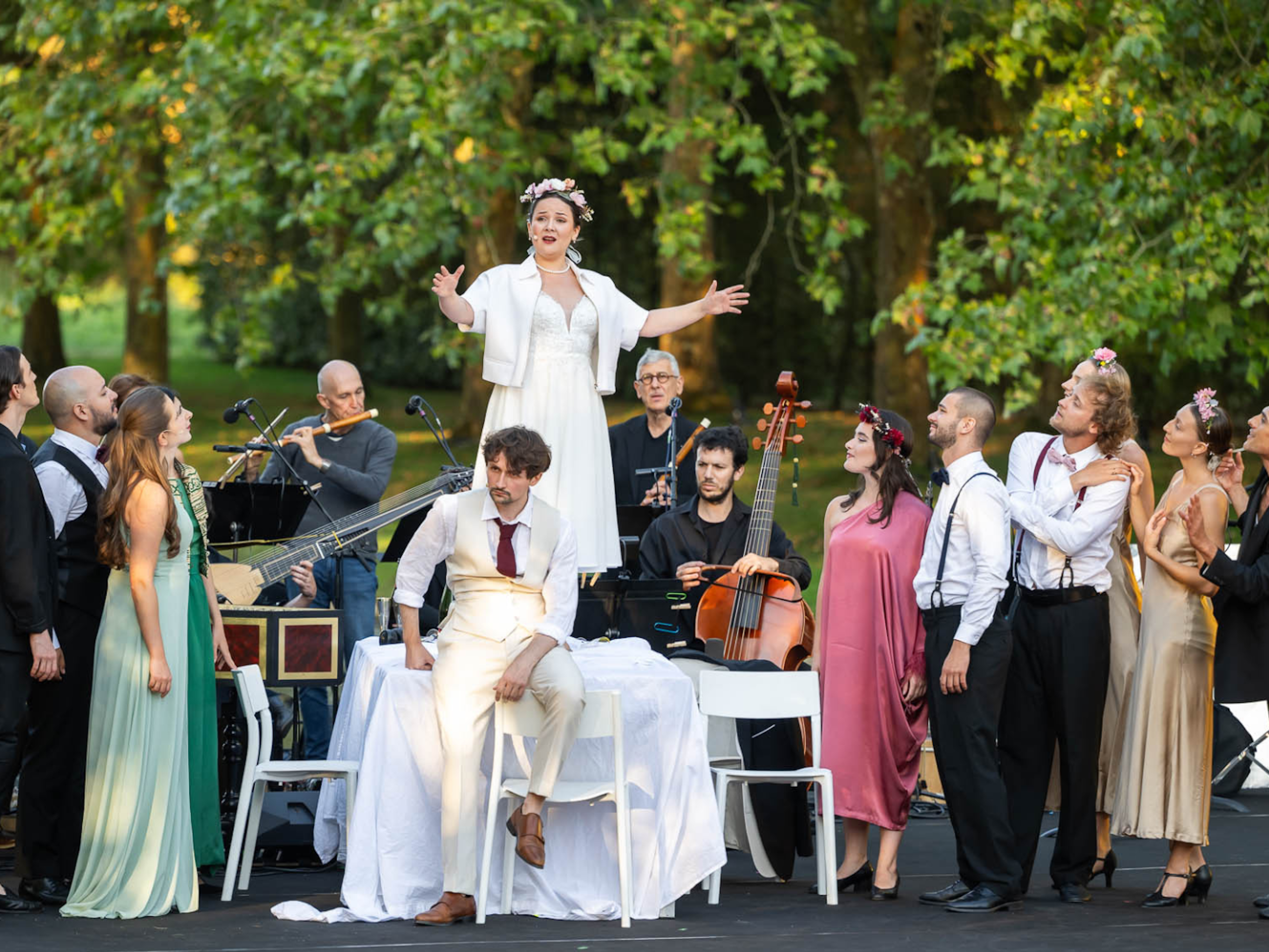 A baroque opera group performs an opera in a lush outdoor setting. A woman stands on a table singing while the opera group, in costume, look up at her.