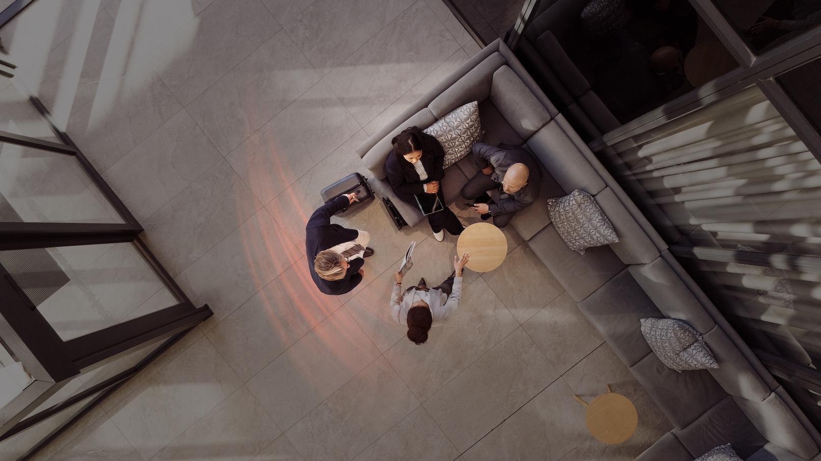 Two people sit on a sofa as they are greeted by a member of staff, a man with a suitcase stands nearby