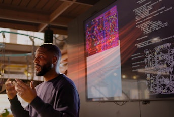 A man speaks while standing infront of a large screen displaying code