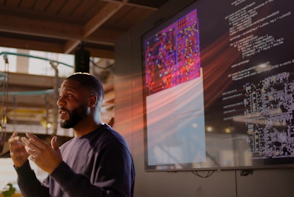 A man speaks while standing infront of a large screen displaying code