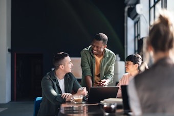 A man and a woman sit at a table with a laptop and coffee, another man is standing at the desk speaking
